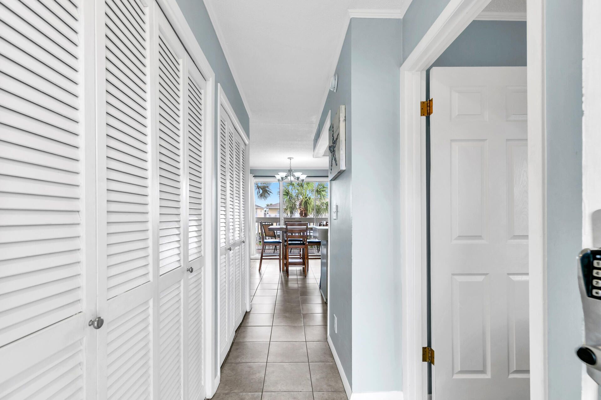 A hallway with white shutters, a dining area with a table and chairs, and a door with a keypad lock.