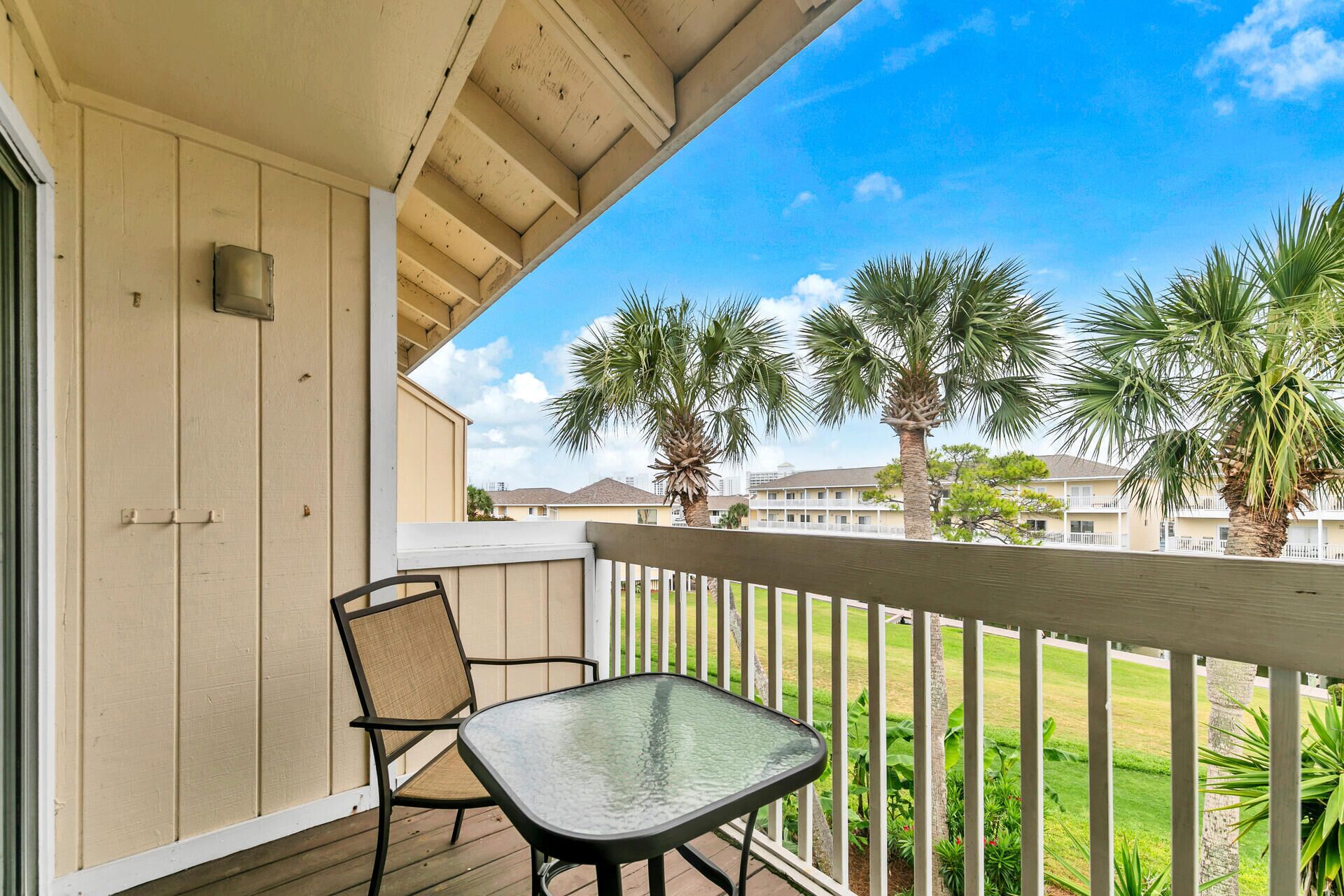 A balcony with a glass table and two chairs, overlooking a lawn with palm trees and a building in the background.
