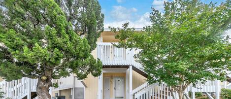 A two-story building with a wooden deck leading to the entrance, surrounded by trees and a white fence.
