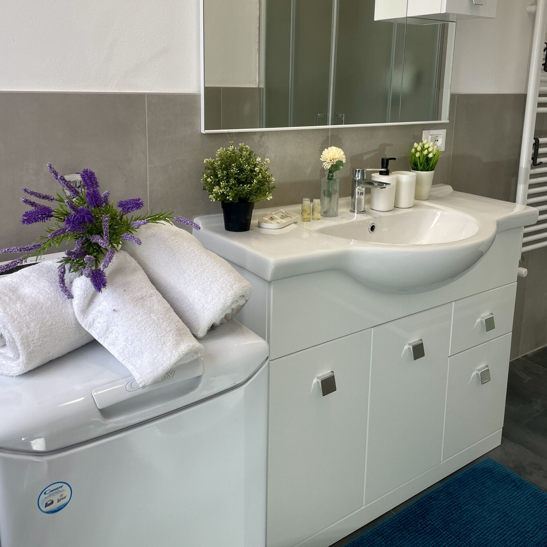 A bathroom with a white vanity, a mirror, and a sink. There are towels, a potted plant, and toiletries on the vanity.