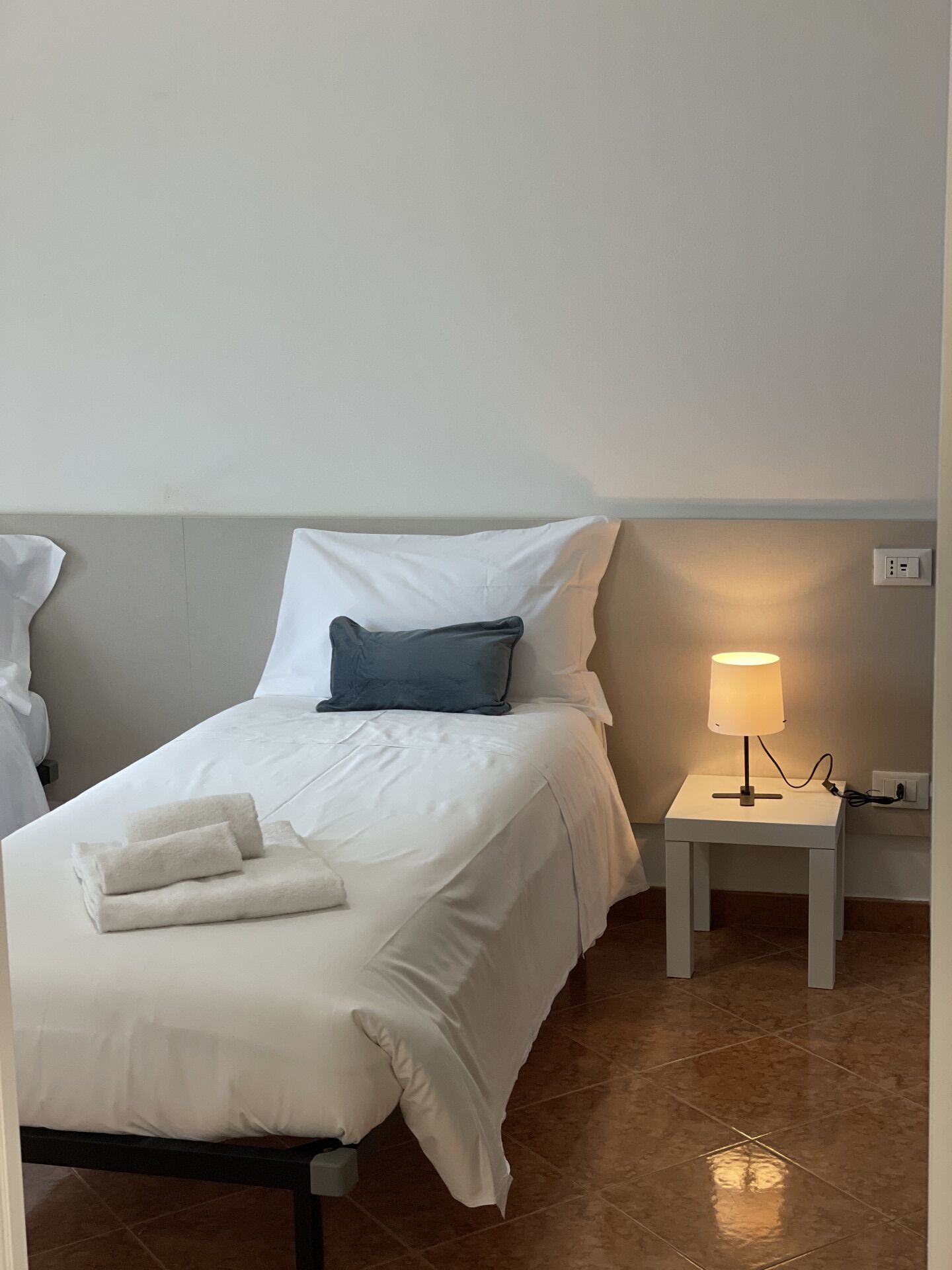 Un lit bien fait, avec du linge de lit blanc et un oreiller bleu, une petite table de chevet avec une lampe, et une robe accrochée au mur.