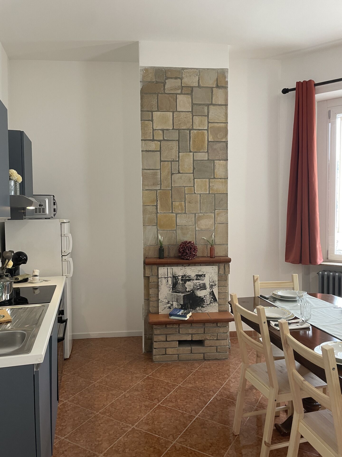 A compact kitchen with a stone backsplash, a dining table with chairs, and a red curtain near a window.