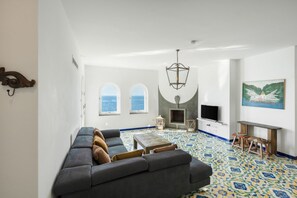 A modern living room with a sofa, a fireplace, a television, and a view of the sea through two windows. - Villa Stunning view on the sea of Naples with private pool and beach access (San Montano)