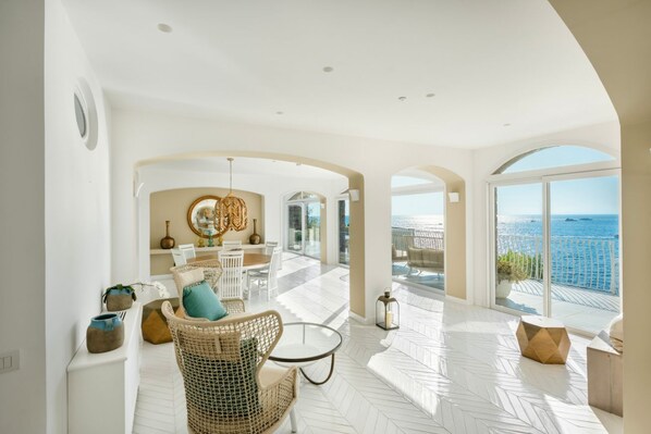 A spacious indoor area with a view of the sea, furnished with wicker chairs and a table, and a ceiling with recessed lighting.
