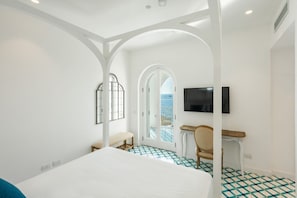 A hotel room with a bed, a desk, a chair, a TV, and a balcony with a view of the sea. - Villa Stunning view on the sea of Naples with private pool and beach access (San Montano)