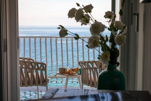 A balcony with a view of the sea, a table with breakfast items, and a vase of flowers.