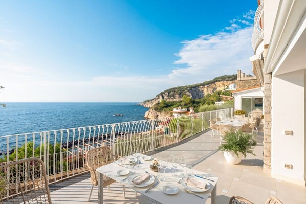 A terrace with a table set for dining, overlooking a coastal landscape with cliffs and the sea.