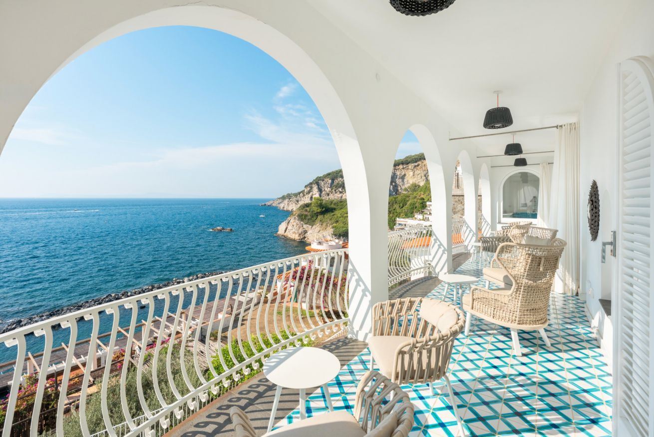 A balcony with a view of the sea, white arches, wicker furniture, and a tiled floor.