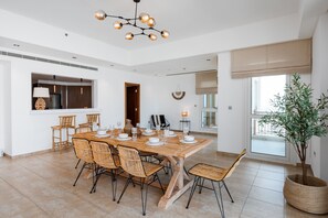 A spacious dining area with a large wooden table set for six, surrounded by wicker chairs. A modern chandelier hangs from the ceiling, and a potted plant is visible to the side.