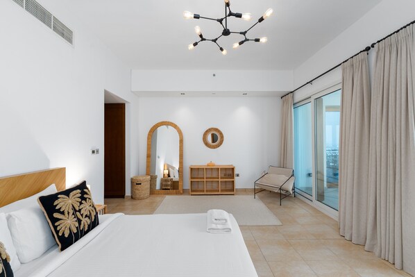 A modern bedroom with a large bed, a wooden headboard, a chandelier, a mirror, a vanity, and a view of the city through large windows.