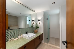 Un baño moderno con un espejo grande, un lavamanos blanco y un salpicadero de azulejos verdes. Hay una planta en maceta en la encimera y se ve una puerta de ducha al fondo.