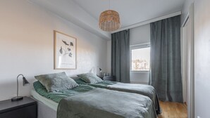 A bedroom with two beds, a bedside table with a lamp, a framed picture on the wall, and a window with curtains.