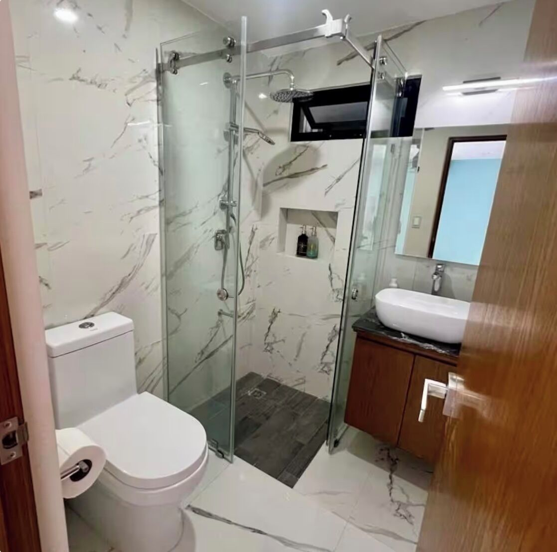Une salle de bain dotée d’une douche à parois de verre, de toilettes blanches, d’un lavabo avec un miroir au-dessus et de murs à motifs de marbre.