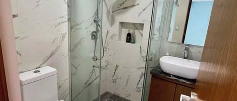 Une salle de bain dotée d’une douche à parois de verre, de toilettes blanches, d’un lavabo avec un miroir au-dessus et de murs à motifs de marbre.