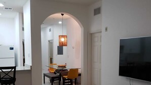 Una sala moderna con un televisor de pantalla plana, un comedor con mesa y sillas, y una cocina con refrigerador.