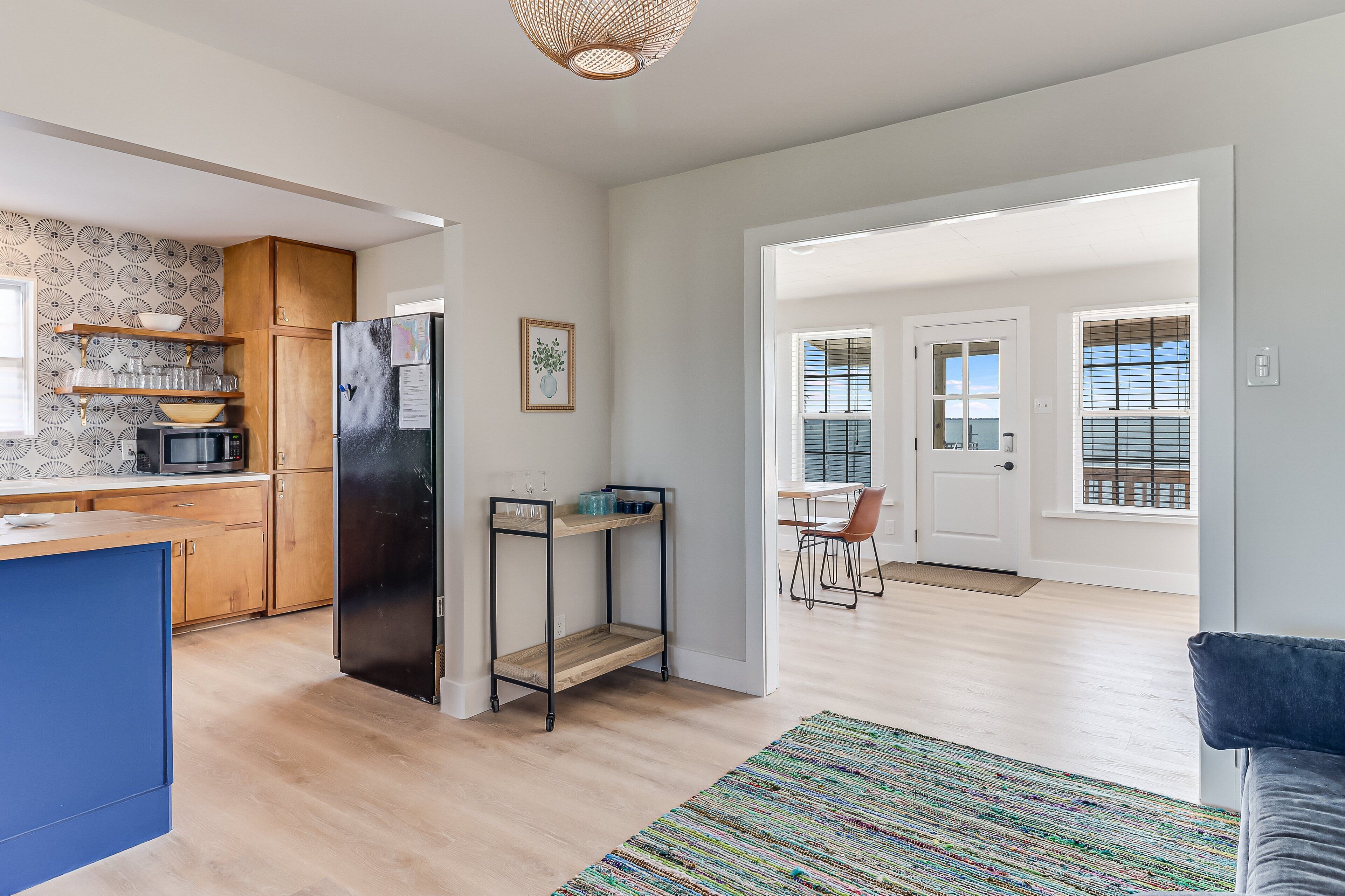 Eine moderne Küche mit Holzausbau, einem schwarzen Kühlschrank und einem Metallregal. Durch eine offene Tür ist ein Essbereich mit Tisch und Stühlen erkennbar.