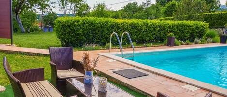 Zona junto a la piscina con mobiliario de mimbre y una mesa de vidrio, rodeada de follaje y un cielo azul despejado.