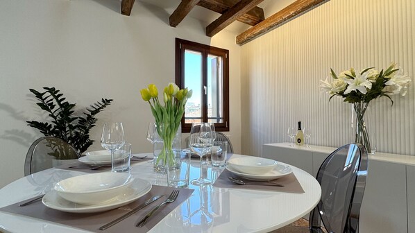 Una mesa de comedor preparada con platos, cubiertos y vasos, con un jarrón de flores y una botella de vino en una mesita auxiliar.