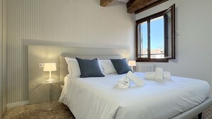 A neatly made bed with white and dark blue pillows, a glass side table with a lamp, and a window with a view of buildings. - Salieri Attic (500mt. from St. Mark's Square) (Venecia)