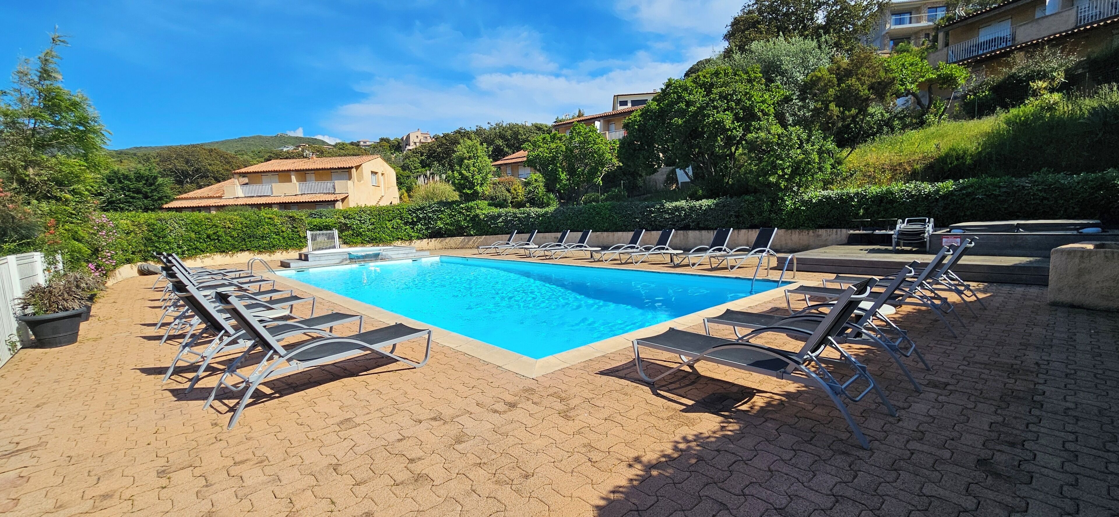 Una piscina rectangular con sillones de descanso alrededor, una zona pavimentada y edificios al fondo.