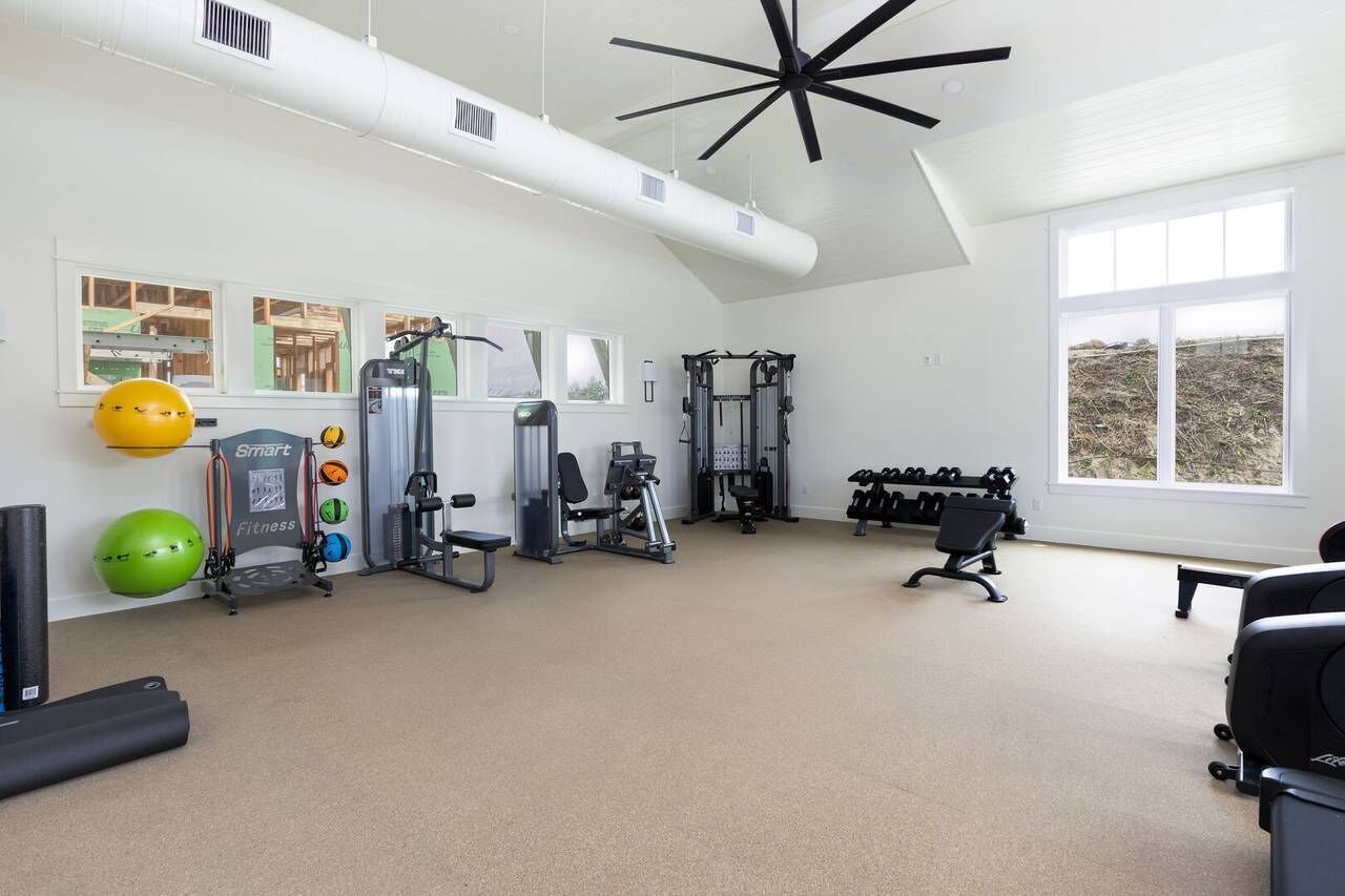 Uma academia ampla com diversos equipamentos de exercício, janelas grandes e um ventilador de teto.