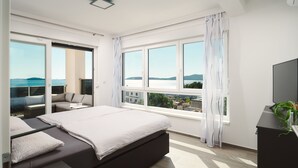 A modern bedroom with a large bed, a TV, and a balcony with a view of the sea.