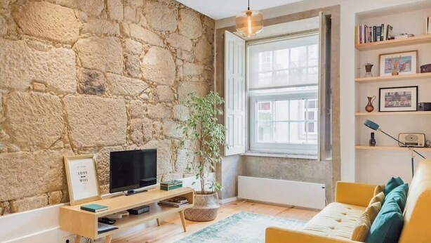 Una sala de estar con pared de piedra, televisor, planta, sofá amarillo y estantería.