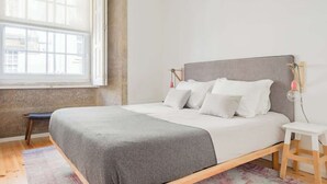 Un dormitorio minimalista con un ventanal grande, una cama con cabecero gris y una mesita de noche pequeña.