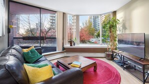 A modern living room with a leather sofa, a coffee table, a TV stand, and a large window offering a city view.