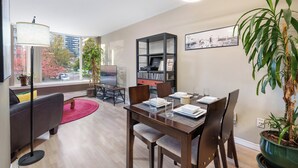 A modern living room with a dining area, large windows, a bookshelf, and a potted plant.