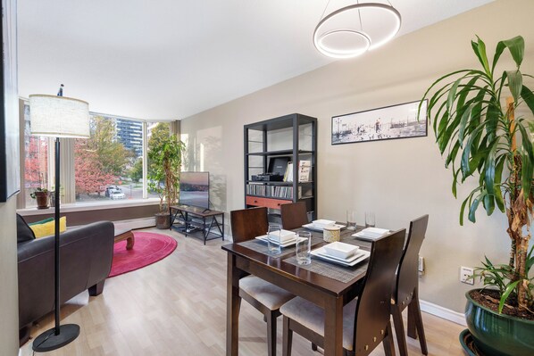 A modern living room with a dining area, large windows, a bookshelf, and a potted plant.