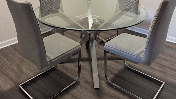 Een ronde glazen tafel met vier stoelen met kussens, een houten vloer en een effen muur.