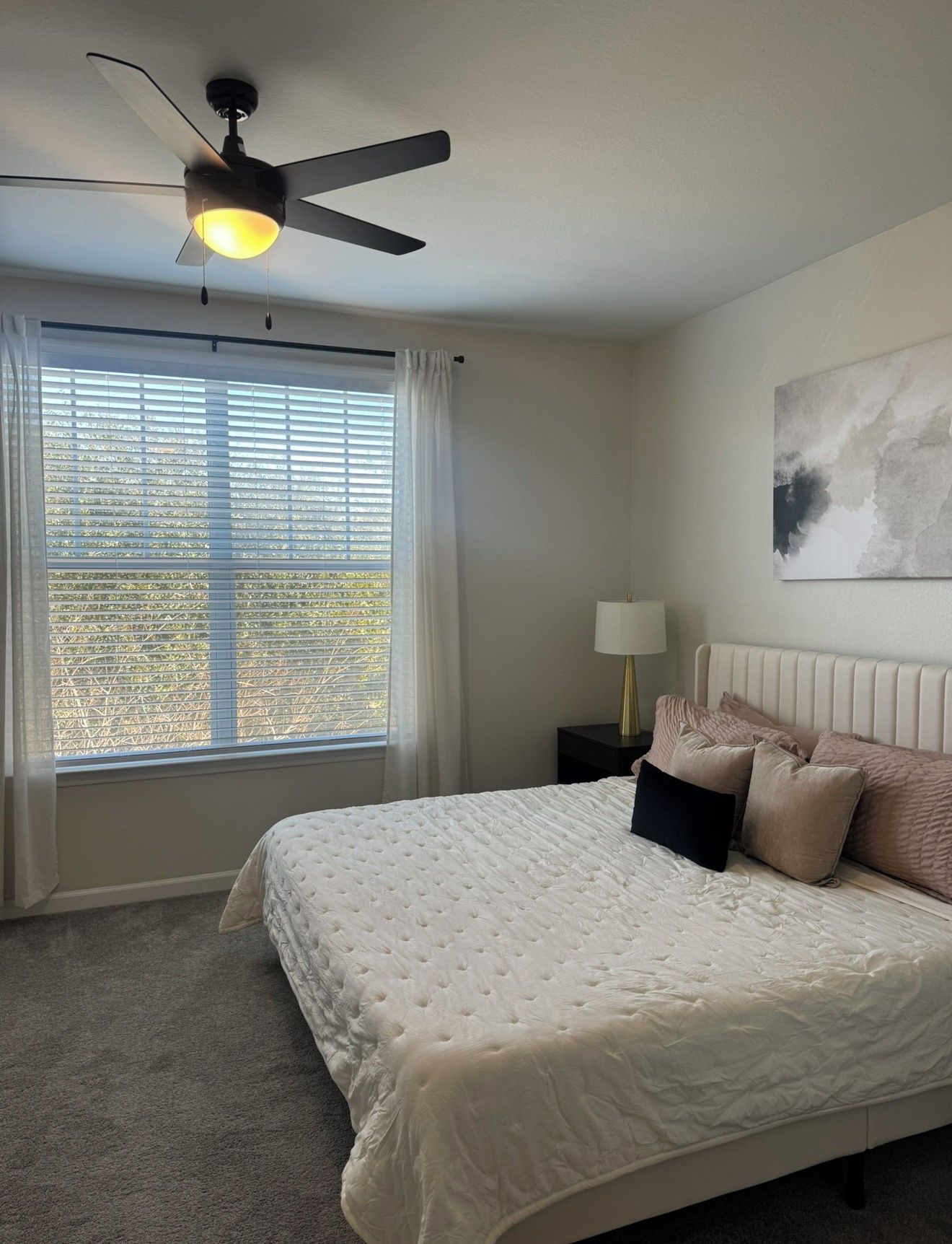 Une chambre à coucher avec un lit, un ventilateur de plafond, une fenêtre avec des stores, une table de chevet avec une lampe et un tableau au mur.