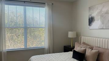 A bedroom with a bed, a ceiling fan, a window with blinds, a nightstand with a lamp, and a painting on the wall.