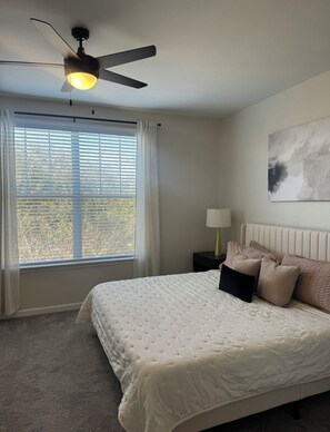 A bedroom with a bed, a ceiling fan, a window with blinds, a nightstand with a lamp, and a painting on the wall. - Heart of Bartram Escape! (Jacksonville)