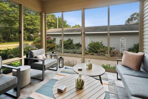 A covered patio with outdoor furniture, a round table, and a view of a garden and house.