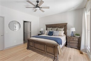 Un dormitorio con una cama de madera, una mesita de noche con una lámpara, un ventilador de techo y un espejo grande en la pared.