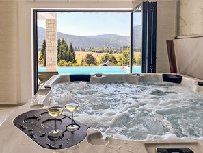 Un bain à remous avec deux verres de vin et une télécommande, donnant sur une piscine et un paysage pittoresque.