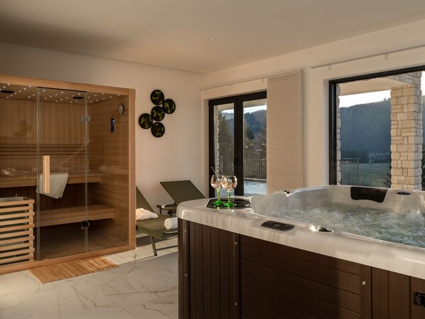 A modern spa room with a hot tub, wooden sauna, and a view of the mountains.