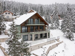 Un chalet moderne à deux étages, doté d’un balcon et entouré d’arbres couverts de neige.