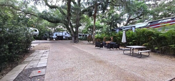 A gravel patio with outdoor furniture, RVs, and lush greenery. - Whispering Oaks Campsite at River Ranch 400 (River Ranch)