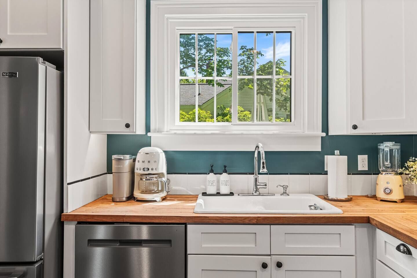 Una cocina con armarios blancos, un refrigerador de acero inoxidable, una cafetera, un lavabo y una licuadora.