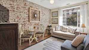 A living room with a brick wall, a sofa, a chair, a table, and a window with curtains. - Above the Tea Room in the Heart of Downtown (Savannah)