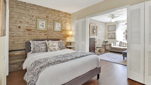 Une chambre avec un mur en briques, un lit recouvert d’une literie à motifs, une table de chevet avec une lampe, et une vue sur une autre pièce où se trouvent un canapé et un ventilateur de plafond.