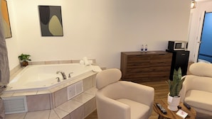 A modern bathroom with a freestanding bathtub, two beige armchairs, a wooden side table, and a small potted plant.