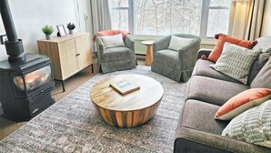 Un salón acogedor con una estufa de leña, dos sillones verdes, un sofá gris y una mesa de centro redonda.
