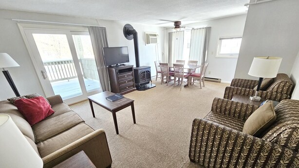 A living room with a sofa, a coffee table, a TV, a fireplace, a dining table with chairs, and a ceiling fan. - Centrally located 2BR condo, Hot Tub&Indoor Pool (Stowe)