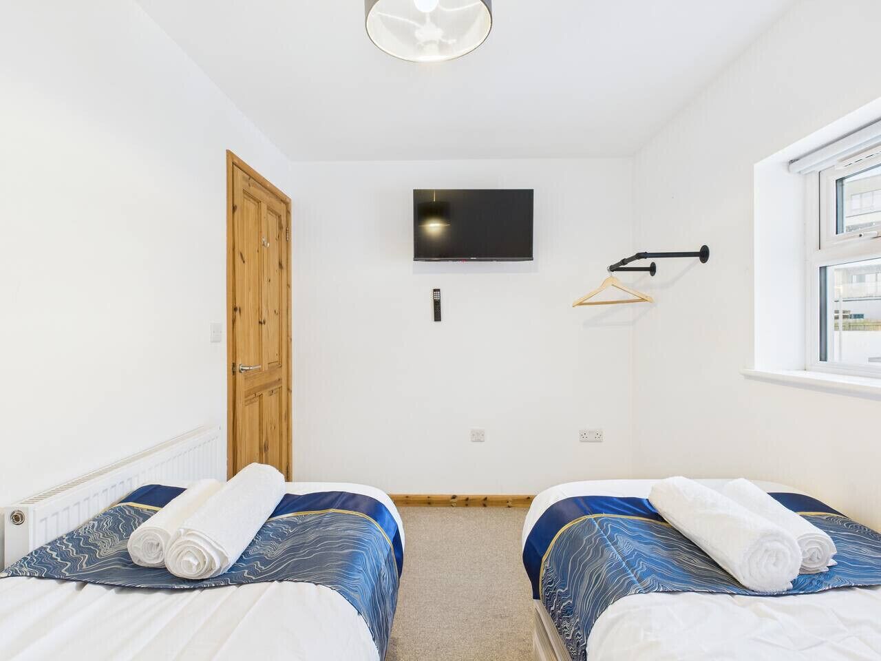 A bedroom with two beds, a TV mounted on the wall, a wooden door, and a window with a view.