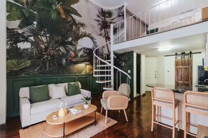 Een moderne woonkamer met een witte bank, een houten salontafel en rieten stoelen. Een spiltrap leidt naar een hoger niveau.