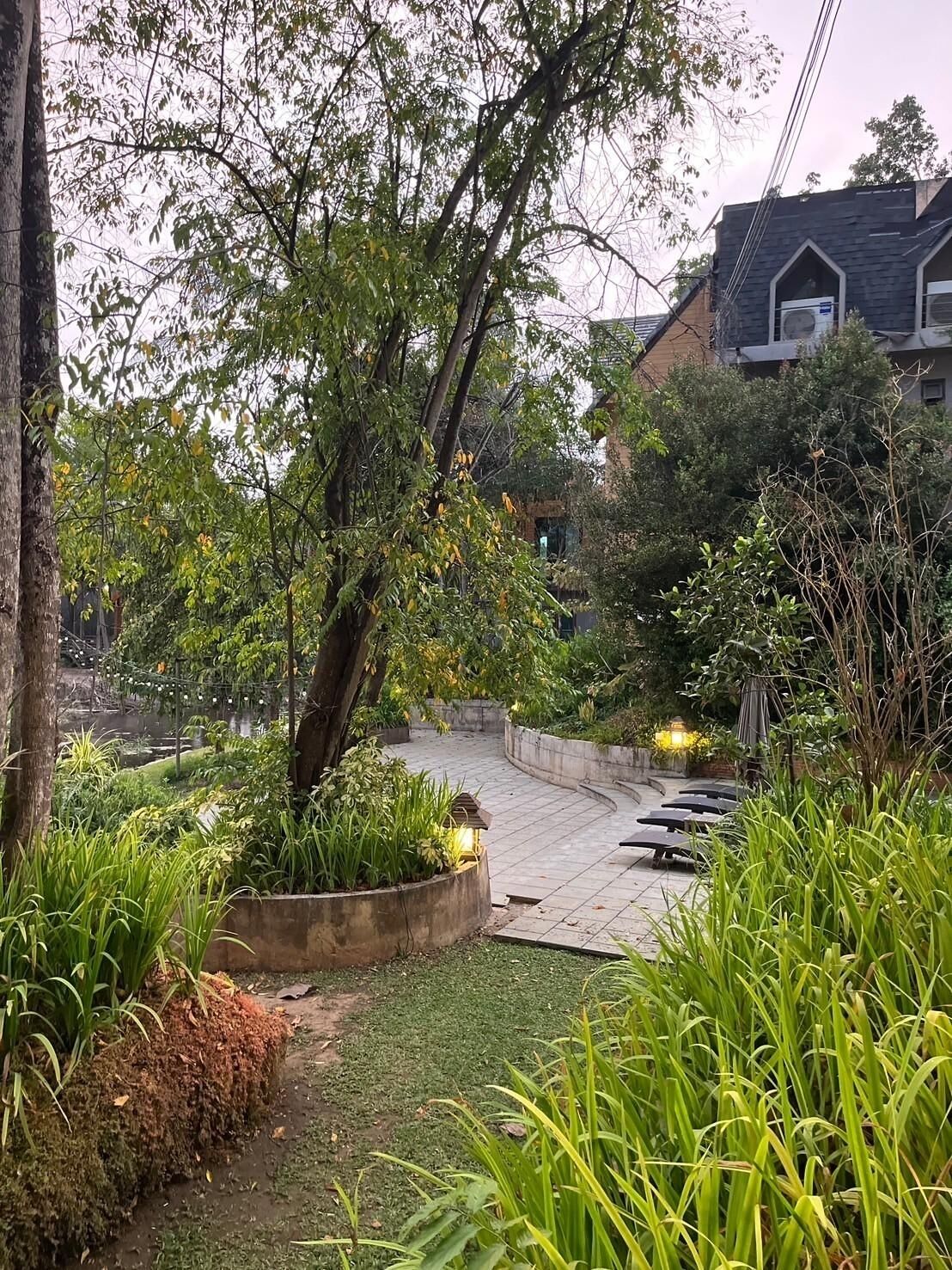 Uma área de jardim com um caminho sinuoso, rodeada por diversas plantas e árvores. Há áreas de estar ao ar livre com mesas e cadeiras.